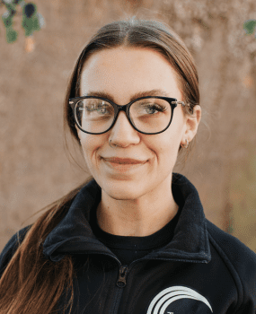 A woman with glasses wearing a black jacket, looking confidently at the camera.